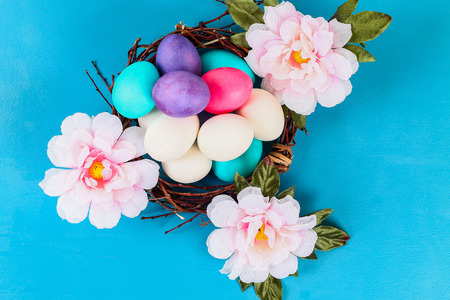Easter colored multicolored eggs lying inside the nest next to the flowers on a blue background....
