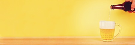 A man pours beer into a mug from a bottle on a yellow background on a wooden table. Copy spaceの写真素材