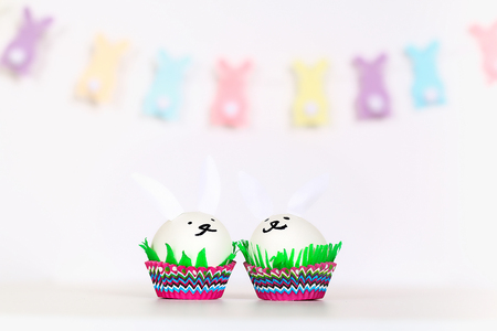 Diy rabbit from easter eggs on white background. Gift ideas, decor Easter, spring. Handmade. White easter egg bunny, grass crepe paper, cup muffin cupcake. kid crafts.の写真素材
