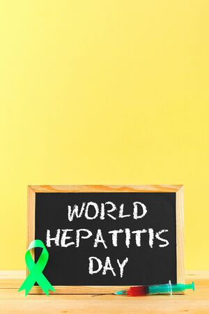 Chalkboard with text World Hepatitis Day. June 28th. A green jade ribbon and a syringe with blood on a wooden table against the background of a yellow wallの写真素材