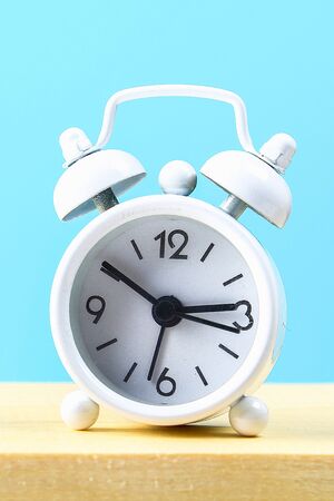 White small alarm clock on a wooden shelf on a blue pastel background. Minimalism.の写真素材