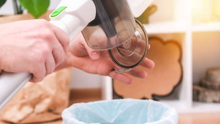 A man shakes out a trash container of a wireless vertical vacuum cleaner into a trash can. The...