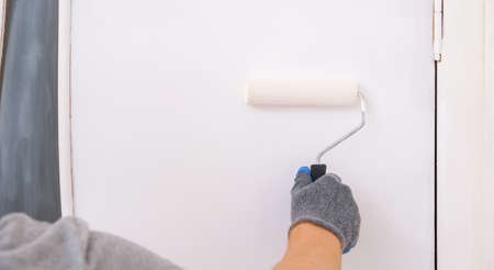 A man paints a wooden door white. The painter uses a foam paint roller to apply white acrylic paint...