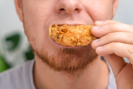 A man bites and chews a fried chicken wing in breadcrumbs. A man eats fast food. Unhealthy diet, bad habits.の写真素材