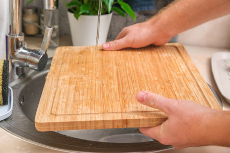 A man washes a wooden bamboo cutting board in the kitchen sink under running water. Gentle hand washing of a wooden cutting board. Cleaning dirty kitchen wood products.の写真素材