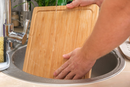 A man washes a wooden bamboo cutting board in the kitchen sink under running water. Gentle hand washing of a wooden cutting board. Cleaning dirty kitchen wood products.の写真素材