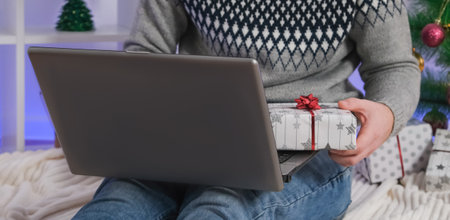 Man with laptop and gift buys gifts for New Year, Christmas. Man in Santa hat on background of Christmas tree, garlands, gifts with laptop. Online shopping, shopping in an online store, marketplace.の写真素材