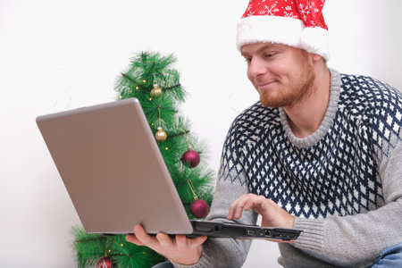 Man with laptop working, studying remotely new year 2023, Christmas. Man in Santa hat on background Christmas tree, garlands, gifts. Remote work. Surfing Internet. Man congratulates on holiday online.の写真素材