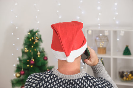 Man in a christmas hat is talking on the phone Christmas Eve or New Years Eve. A young man in a hat makes greetings phone for Christmas. Congratulations your loved ones Christmas by phone.の写真素材