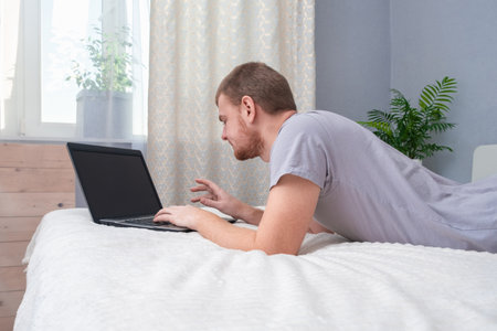 A man with laptop is lying in bed. Freelancer works remotely from home. Distance education. Online shopping without leaving home. Internet surfing. Gambling addiction. Black Friday sale. Cyber Monday.の写真素材