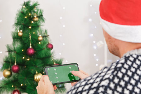 A man in Santa hat holds a phone in a horizontal position with a green screen. Phone with chromakey. Copy space. The concept of advertising, New Years holidays, Christmas.の写真素材