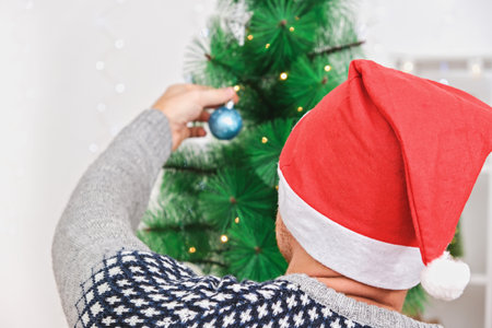 Preparation for the new year. New Year and Christmas decor. A man decorates a tree with toys. Preparing for Christmas. A man in a Christmas hat decorates a Christmas tree for the holiday.の写真素材