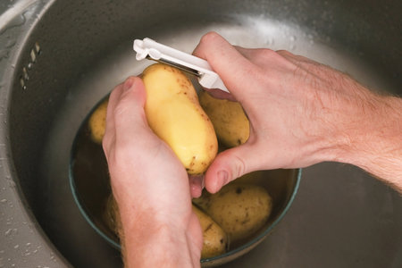 A man peels potatoes with a vegetable peeler in the sink. Preparation of potatoes for cooking. Cleaning the peel from pesticides. A man cooks dinner or lunch at home for the family.の写真素材
