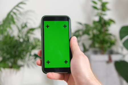 A man holds a phone with a green screen in his hands against a background of green house plants. mockup. Chroma key.の写真素材