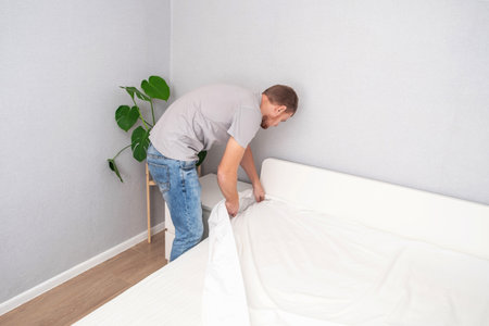 A man makes his cozy bed with fresh bright white bed linen. Making the bed with fresh bed linen by a white man. The day of the change of bedclothes. The day of washing bedclothes in the laundry room.の写真素材
