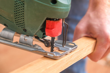 The hands of a master carpenter with an electric jigsaw in his hands cutting off a piece of wood. Male hands using fret saw for cutting wood. A carpenter cuts wood with a jigsaw in a home workshop.の写真素材