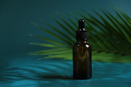 Drop of clear liquid, oil, serum or tincture dripping from pipette in the neck of a brown medical bottle on a turquoise background with palm leaves.の写真素材