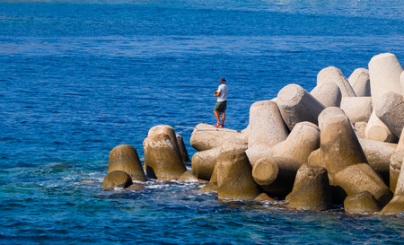 Man fishing, standing on huge concrete tetrapods - blue sea and gray concreteの写真素材