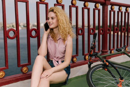 Young woman with bike sitting on a sideways of a bridge near to water and calling by phone. Girl...