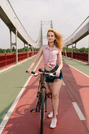 Urban biking. Young woman with bike on a bicycle road. Pretty woman in a jeans shorts near the bike...