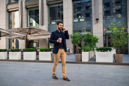 Confident young manager walking outdoors in front of cafe talking by phone and holding coffeeの写真素材