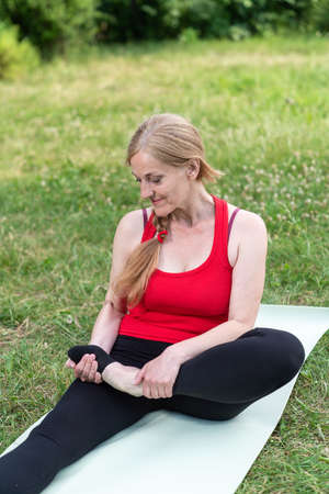 50 years old woman stretching on mat outdoors in a park on a grass.の写真素材