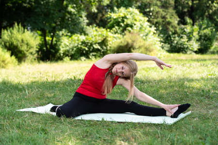 50 years old woman practice yoga on mat and stretching outdoors in a park on a grass. Woman in red topの写真素材