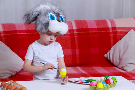 Little boy paints eggs with colored paints in a bunny cap