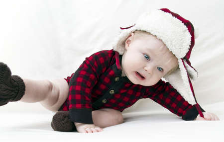 Surprised Christmas baby with red plaid shirt and furry wool hat falling over while trying to pull off capの写真素材