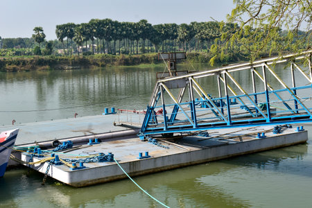 This image features a metallic blue floating pontoon bridge or ferry dock on a calm river, with a ramp for vehicles or pedestrians. Lush greenery and tall palm trees line the riverbank, creating a picturesque contrast between industrial elements and natural beauty.の写真素材