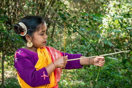 An Indigenous young girl in a vibrant traditional outfit with golden earrings and a pearl hair accessory stands in a lush green forest, holding a handmade bamboo bow and arrow. Her expression reflects determination and curiosity.の写真素材