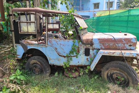 Weathered by time, a vintage jeep or van left in a neglected condition rests under the sun, embraced by nature. Rust and peeling blue paint hint at past adventures.の写真素材