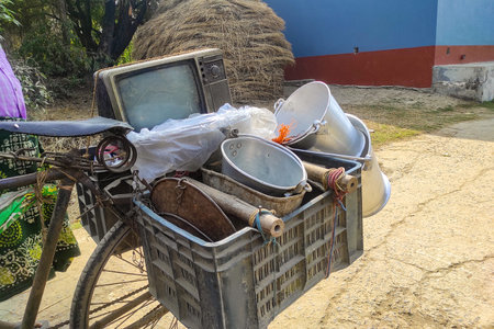 A rural scrap collector transports recyclable household items, vintage CRT televisions, and metal cooking pots on a bicycle. Highlighting economy, recycling and waste collection in developing regions.の写真素材
