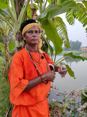 A serene Indian folk singer, dressed in an orange robe, stands by a village backdrop, holding a local string instrument, a peaceful portrait of devotion, tradition, and spiritual calm in rural India.の写真素材
