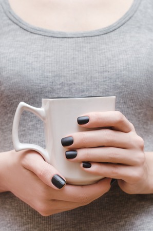 Female hands with black matte nail design holding white cup.の写真素材