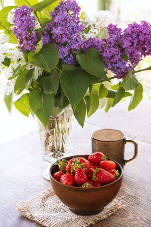Bouquet of lilac branches in crystal vase, clay bowl with red strawberry and dark glass cup on wooden tableの写真素材