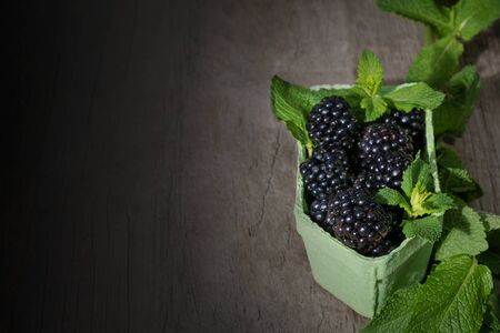 Ripe blackberry berries in a green paper basket with fresh mint leafs on a wooden table.の写真素材