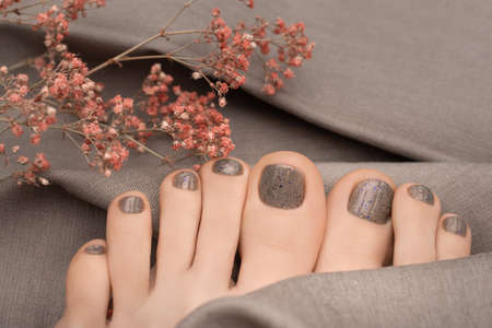 Female feet with beige nail design. Brown nail polish pedicure on gray fabric background