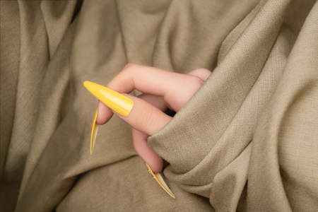 Female hand with yellow nail design. Long nail polish manicure. Woman hand on beige fabric...