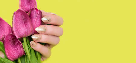 Female hand with golden nail design. Glitter gold nail polish manicure. Woman hand hold pink spring tulips on yellow background. copy spaceの写真素材