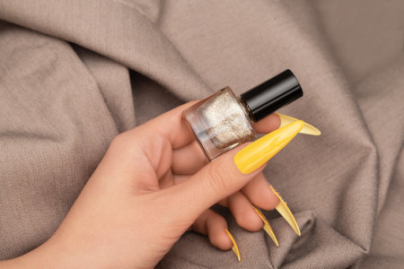 Female hand with yellow nail design. Yellow nail polish manicure. Woman hand hold golden nail polish bottle on beige fabric background.の写真素材