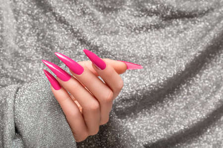 Female hand with pink stiletto nail design. Long nail polish manicure. Woman hand on glitter silver fabric background.の写真素材