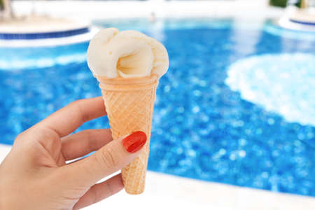 Female hand with red manicure holds yellow ice cream on a hot day on background of a blue pool. The...