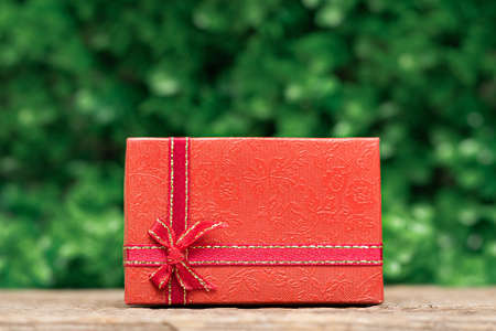 Red gift box with a red ribbon on wooden desk with a green leaves background. Celebration gift cardの写真素材