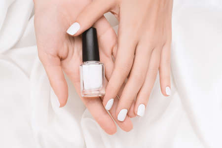 Female hands with white nail design. White nail polish manicure. Woman hands hold white nail polish bottle on white fabric background.の写真素材