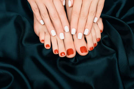 Female hands with white nail design. Glitter red nail polish pedicure. Female hands and feet on dark blue fabric background.の写真素材