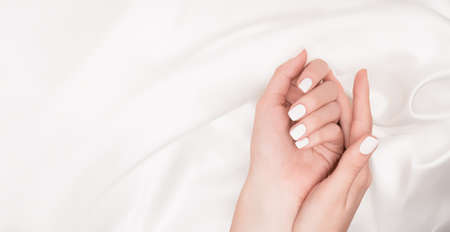 Female hands with white nail design. White nail polish manicure. Woman hands on white fabric background. copy-space. Nails theme template.の写真素材