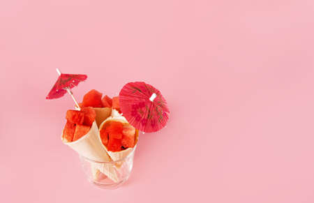 Delicious watermelon cubes in waffle cones on pink background. Red juicy watermelon with red umbrella decoration served in transparent glass. copy space.の写真素材