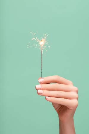 New Year lighted sparkler in female hand on green background. Woman hold glowing holiday sparkling fireworks, shining fire flame. Christmas light.の写真素材