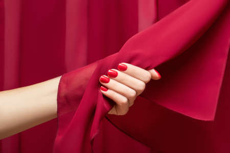 Female hand with red nail design. Glitter red nail polish manicure. Woman hand hold red fabric on red fabric background.の写真素材
